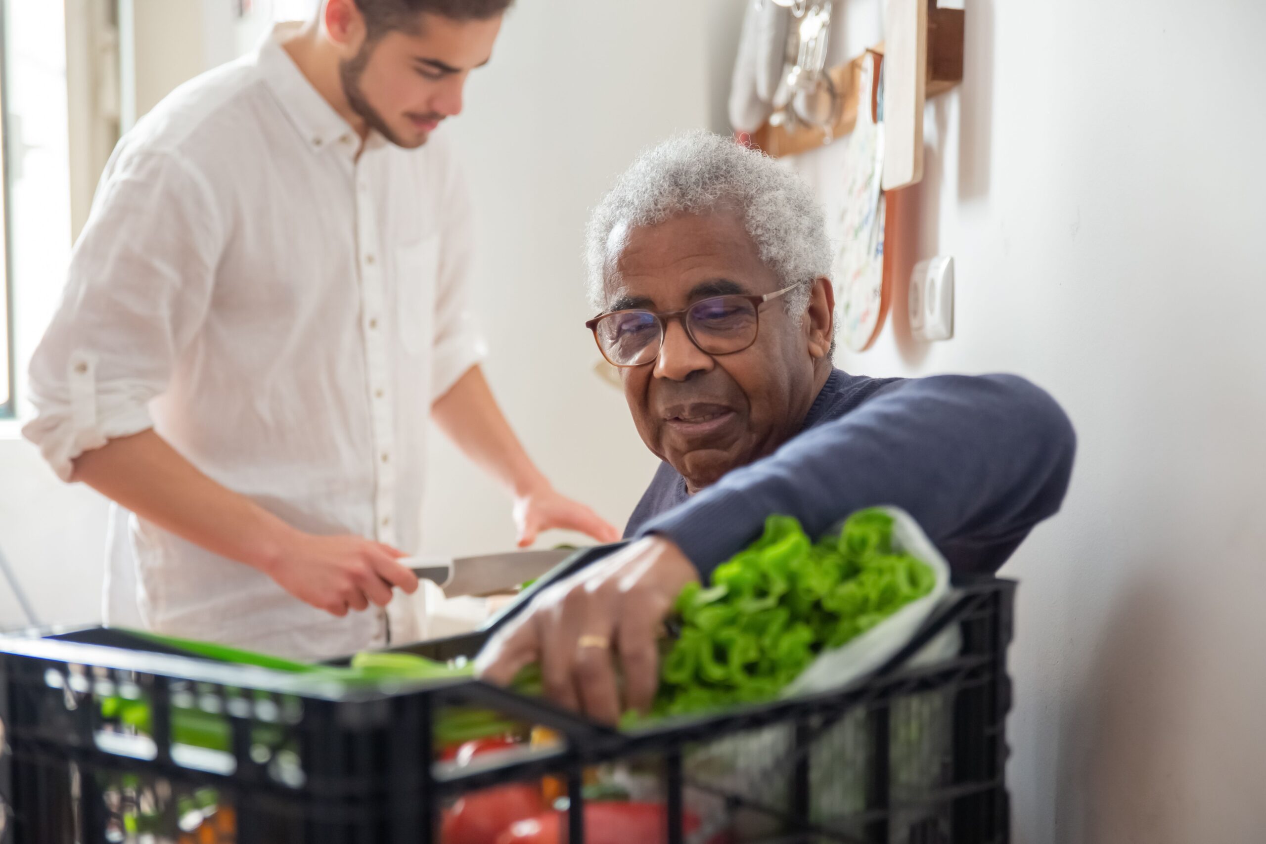 A support worker helping an older man unpack a box of fresh vegetables in his kitchen